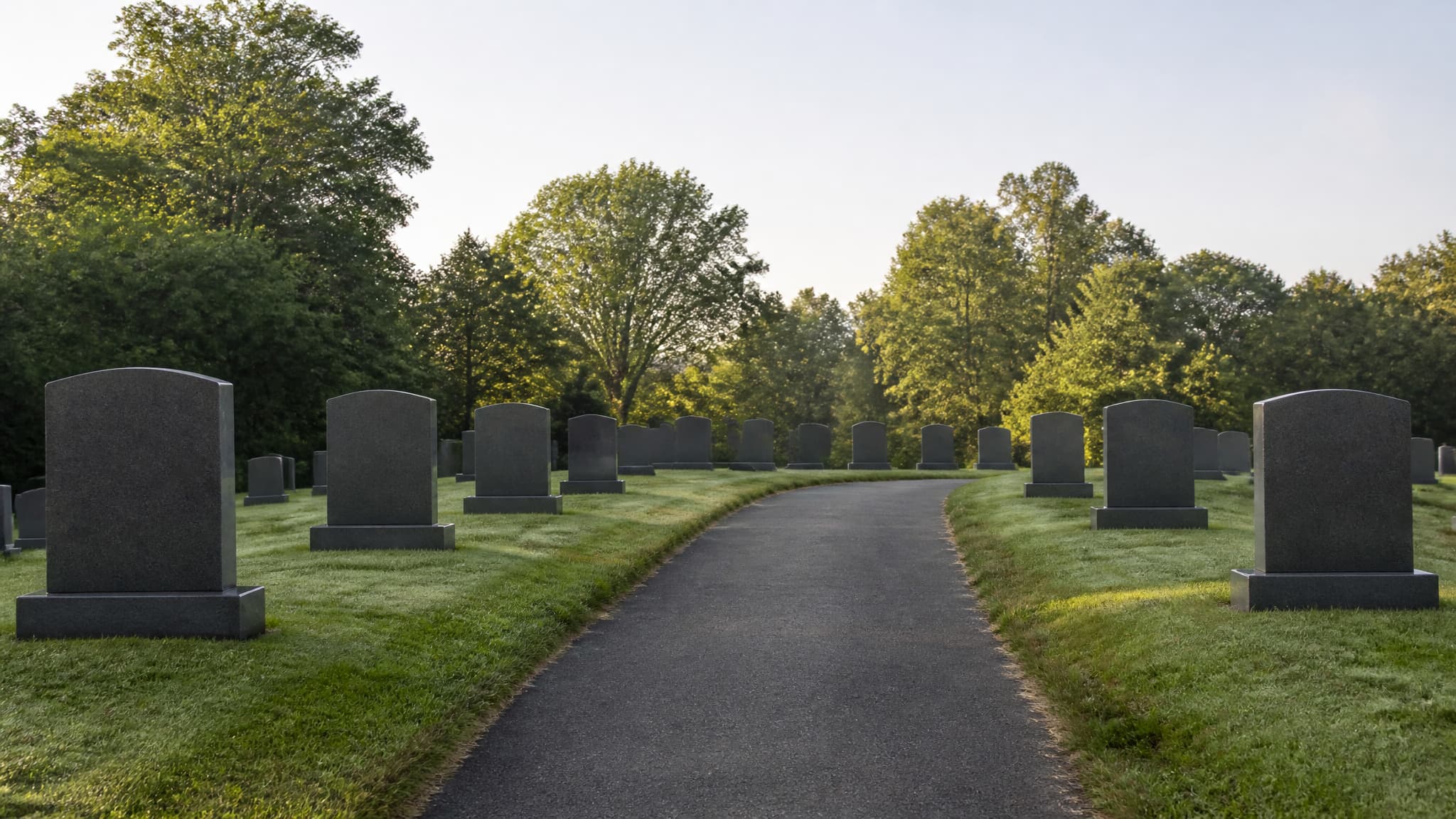 A cemetery path at morning light with granite headstones set along a mown lawn and mature trees behind