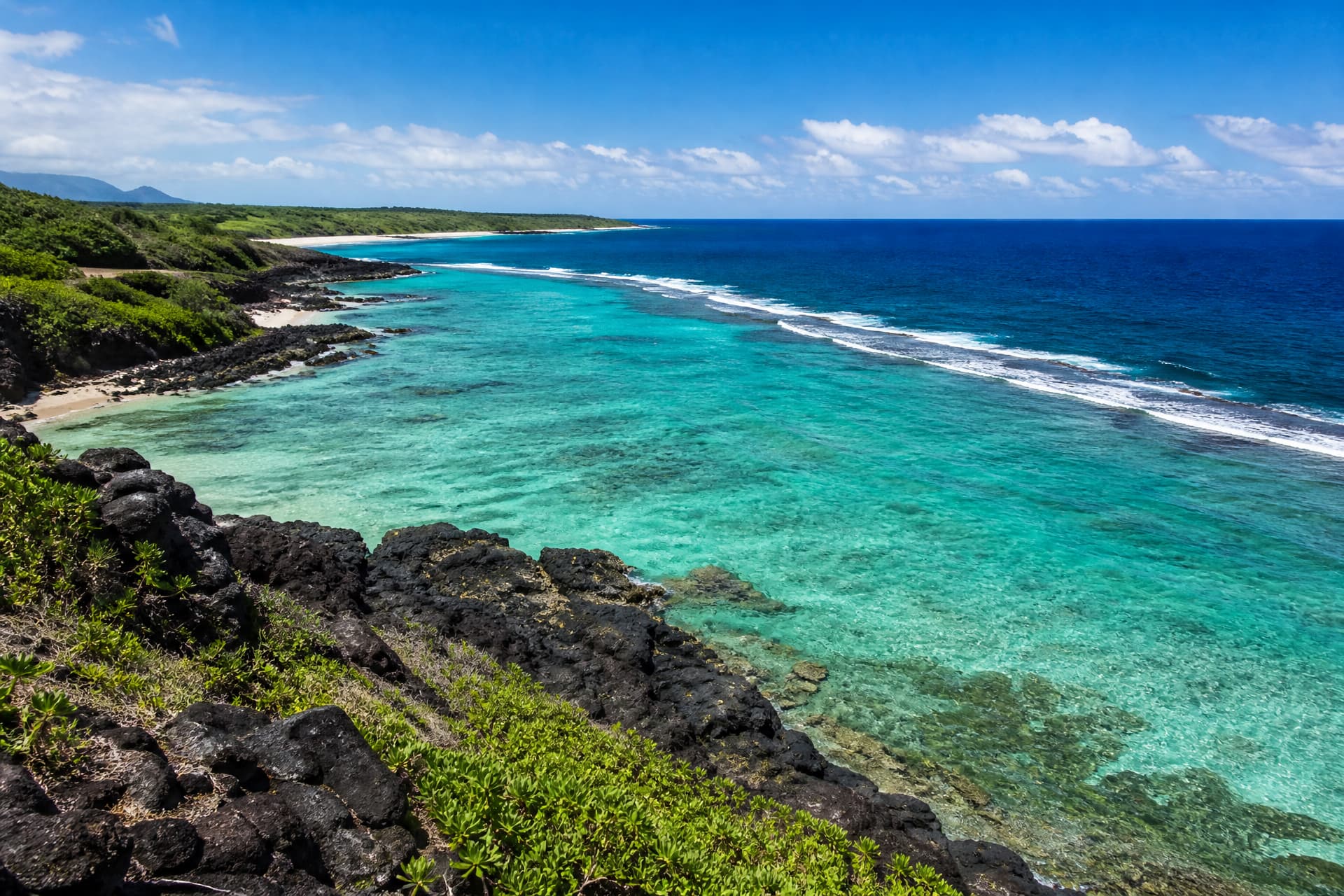 An elevated view along a Hawaiian shoreline in bright mid-morning light showing the pale turquoise shallow water inside the reef, the darker line of the reef itself, and deep blue open ocean beyond it