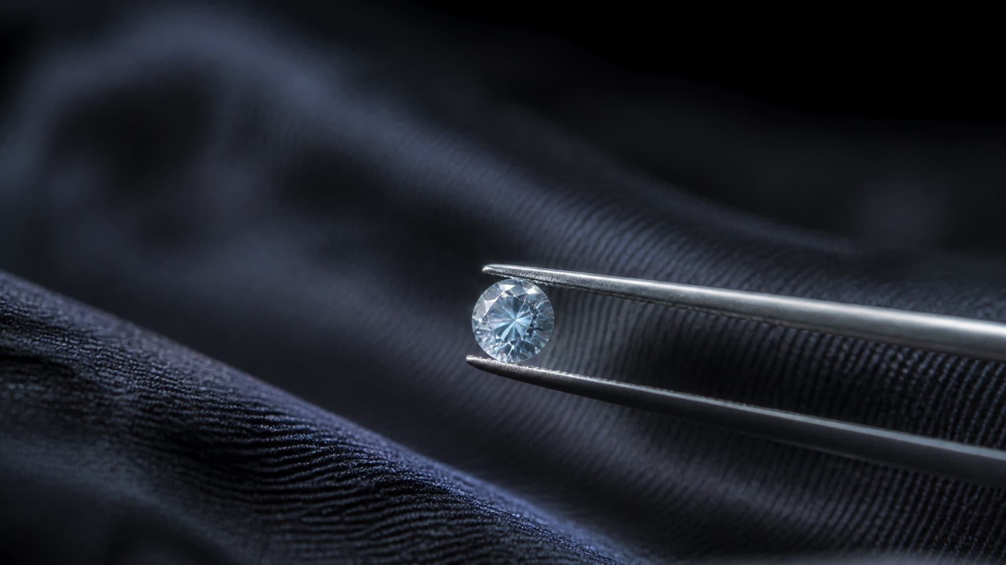 A small pale blue cut diamond held in fine tweezers over a dark cloth under a jeweler's lamp, the facets catching a single point of light